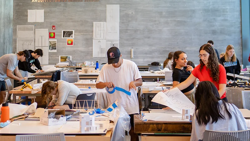 Est&uacute;dio universit&aacute;rio de arquitetura com estudantes trabalhando em maquetes f&iacute;sicas e desenhos sobre as bancadas