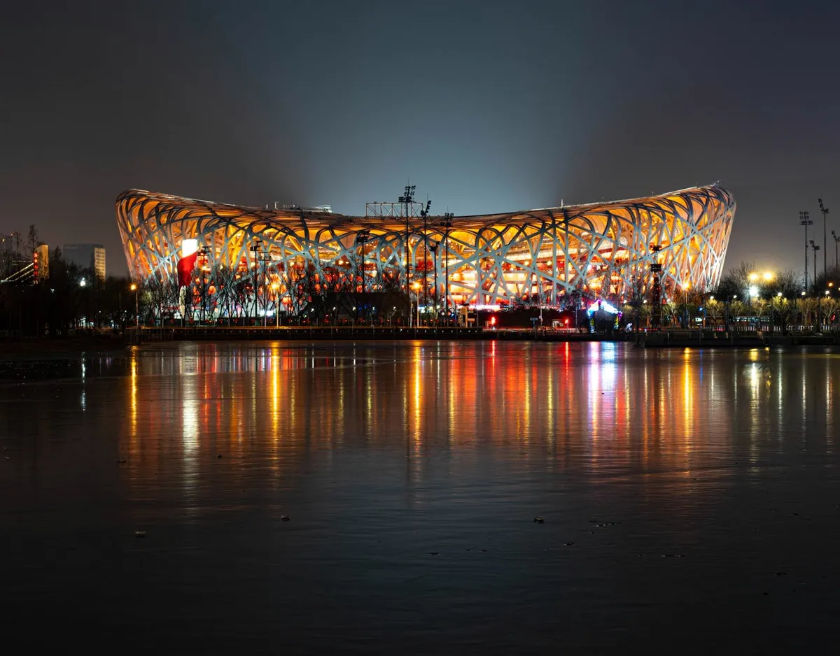 Estádio Nacional de Pequim (Ninho de Pássaro) iluminado à noite, com estrutura de aço entrelaçado refletida na água