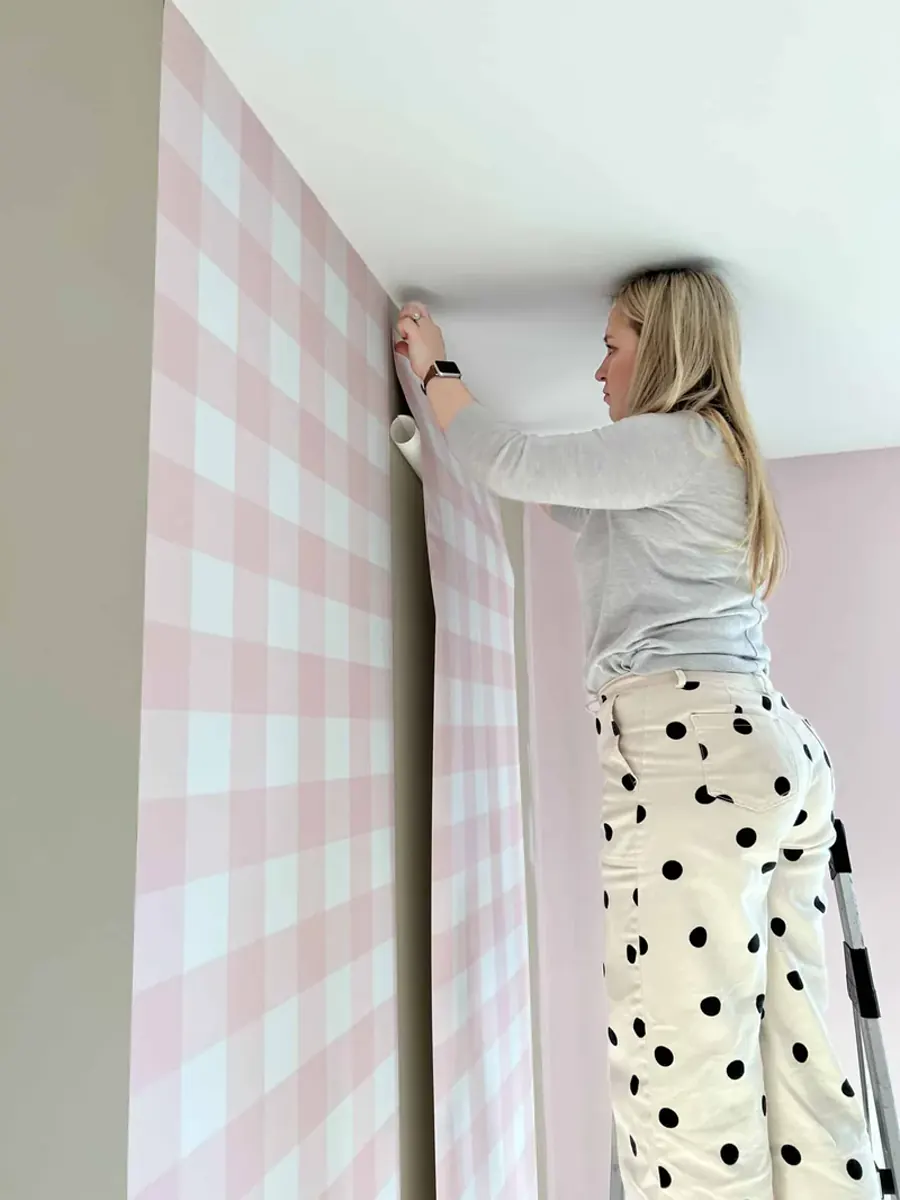 Mulher aplicando rolo de papel de parede xadrez rosa em parede, no canto superior pr&oacute;ximo ao teto