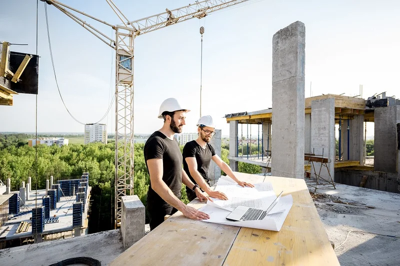 Dois engenheiros analisando planta estrutural impressa sobre prancheta em canteiro de obras com pilar de concreto aparente ao fundo