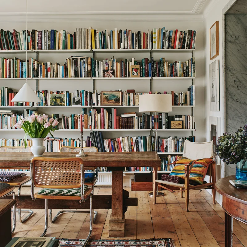 Estante de arquitetura repleta de livros do chão ao teto, com mesa de leitura e cadeiras em sala de luz natural
