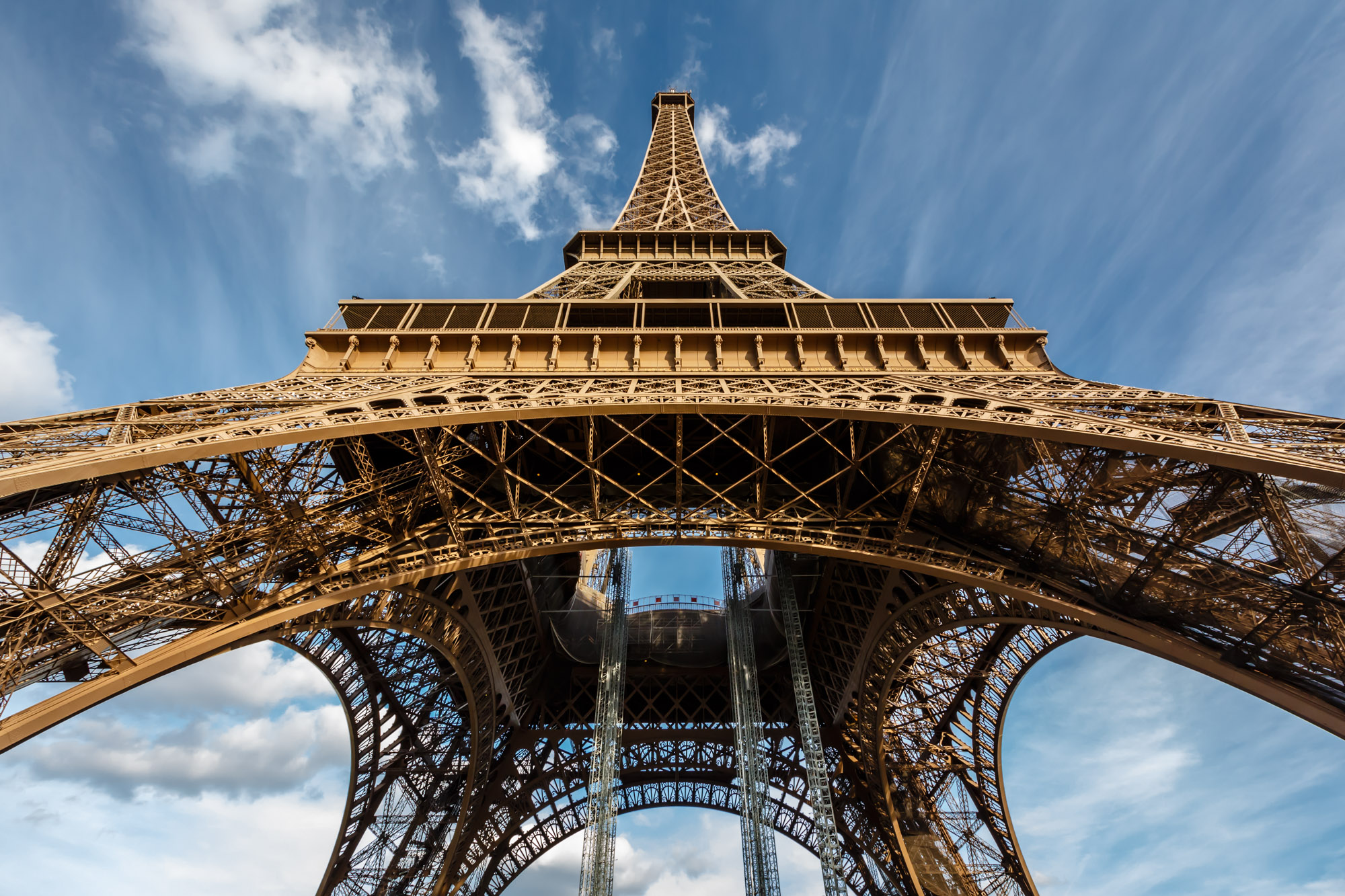 Torre Eiffel vista de baixo contra o céu: os quatro pilares, os arcos e toda a estrutura treliçada de ferro forjado