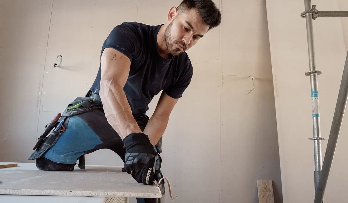 Profissional cortando chapa de gesso acartonado drywall com estilete em canteiro, chapas e perfis metalicos ao fundo