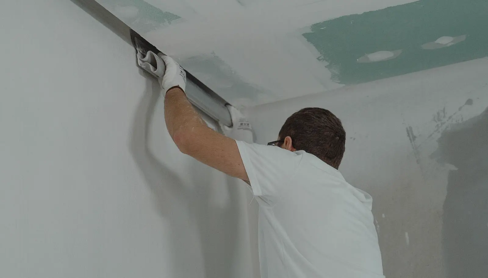 Aplicacao real de drywall: pedreiro alisando junta na transicao parede-teto, chapas RU verdes visiveis no forro recem-fechado